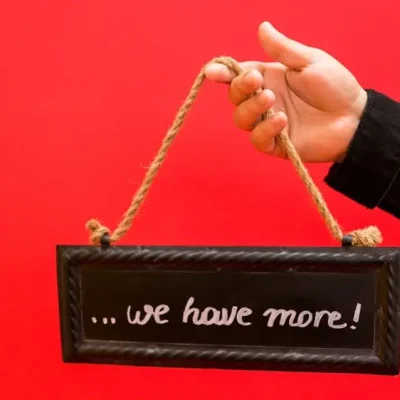 A hand holding a chalkboard sign with 'we have more' against a bright red background.